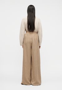 Γυναίκα με μακριά σκούρα μαλλιά, φοράει μπεζ πουλόβερ και ριγέ καφέ παντελόνια με φαρδιά μπατζάκια και ψηλή μέση, στέκεται με την πλάτη γυρισμένη.