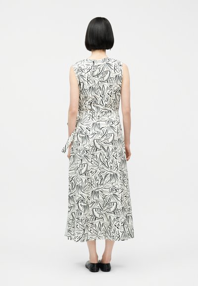 Γυναίκα με κοντά μαύρα μαλλιά, ντυμένη με μακρύ, άσπρο φόρεμα χωρίς μανίκια με αφηρημένα μαύρα φύλλα, από πίσω. Μαύρα παπούτσια slip-on.