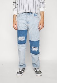 Vaqueros de mezclilla azul claro con acentos de parches azul oscuro, que presentan zonas desgastadas y un logo bordado en la parte frontal. Ajuste casual con una textura sutil.