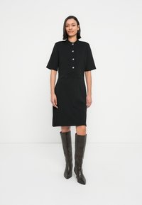 Μαύρο φόρεμα με γιακά, κοντά μανίκια και εφαρμοστή μέση. Εντυπωσιακά χρυσά κουμπιά στο μπροστινό μέρος. Συνδυασμένο με καφέ σκούρες μπότες μέχρι το γόνατο.