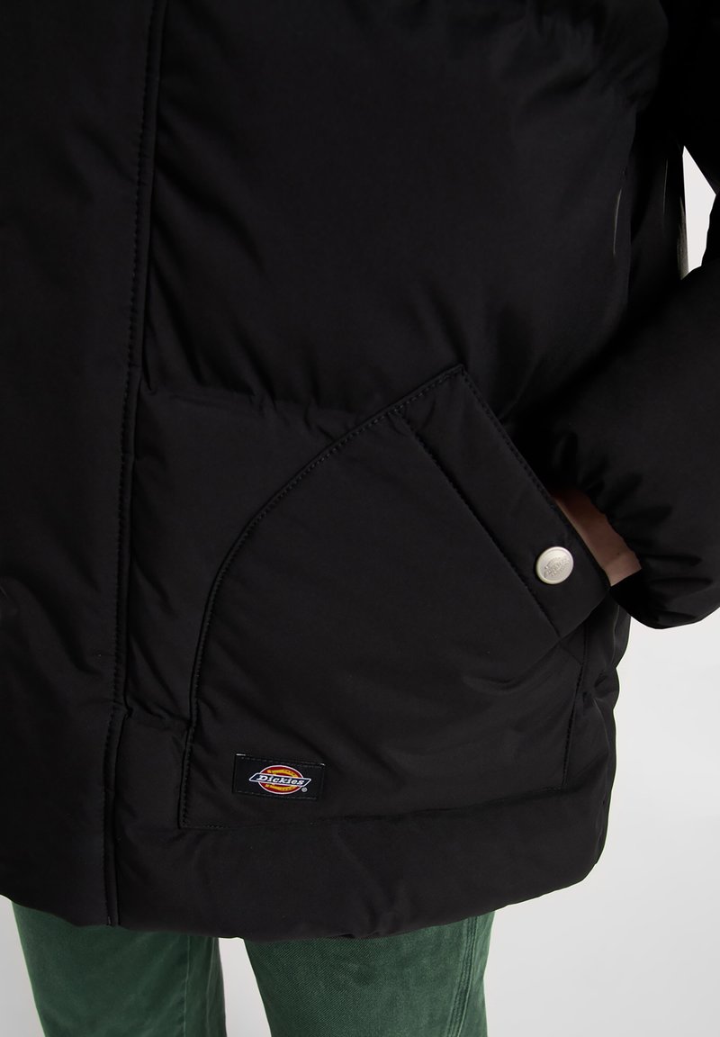 Chaqueta de plumas negra con un gran bolsillo de solapa con botón a presión, detalles de costura y un parche con el logo en el dobladillo. Lleva encima de unos pantalones verdes.