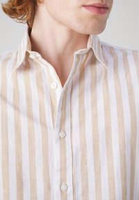 Koszula na guziki w jasnobeżowe i białe paski. Wykonana z lnu, z klasycznym kołnierzem i plastikowymi guzikami na przedniej listwie.