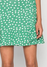 Vihreä pilkullinen mekko, jossa on röyhelöhelma, valmistettu pehmeästä kankaasta; mekossasa on suuria valkoisia pilkkuja tasaisesti jakautuneina koko designin ympäri.