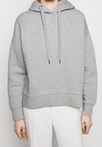 Persoon in een lichtgrijze hoodie met trekkoorden en witte broek, staande tegen een effen witte achtergrond.