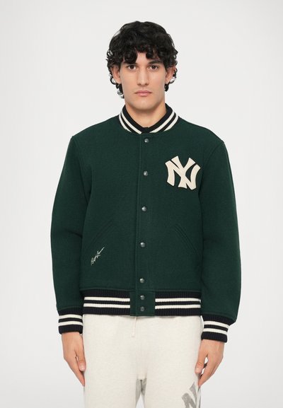 Tânăr îmbrăcat cu o jachetă varsity verde închis, cu guler și mansete albe cu dungi, și pantaloni albi cu un logo pe coapsa stângă, stând nemișcat.