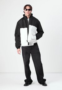 Black and white windbreaker jacket with zip closure, high collar, and front pockets; paired with dark jeans and black shoes.