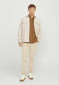 Chemise beige clair à boutons sur une chemise marron, associée à un pantalon en corduroy écru et des baskets blanches avec des semelles beiges. Textures lisses, coupe décontractée.