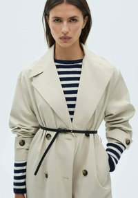 Trench coat beige con vita cinturata, dotato di grandi bottoni e ampi revers. Abbinato a un maglione a righe blu navy e bianche.