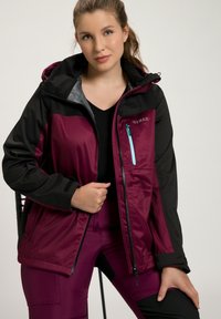 Röd- och svart vattentät jacka med huva, som har en framzip, bröstficka med turkos dragkedja och elastiska ärmslut.