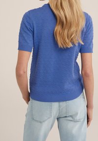 Blå kortärmad tröja med texturerad bobble-mönster, ribbade ärmslut och fåll. Kombinerad med ljusblå jeans, sedd bakifrån.