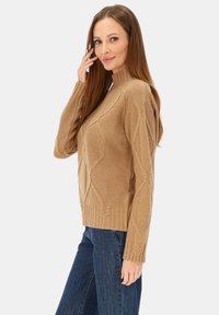 Sweter dziergany w kolorze beżowym, z wysokim kołnierzem, prążkowanymi mankietami i geometrycznymi wzorami warkoczy. W zestawie z niebieskimi dżinsami.