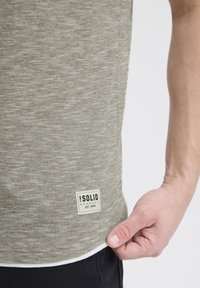 Tricou cu mânecă scurtă, de culoare verde măsliniu, cu dungi subtile, din material de bumbac, având un eticheta albă la tiv cu numele brandului și anul înființării.