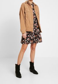 Femme portant une veste en velours côtelé beige sur une courte robe noire à motifs floraux, accompagnée de bottines en suede noires, sur un fond blanc.