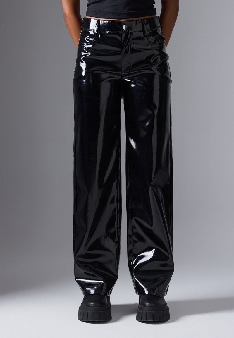 Personne portant un pantalon large noir brillant et des chaussures à plateforme noires épaisses, debout devant un fond gris uni.