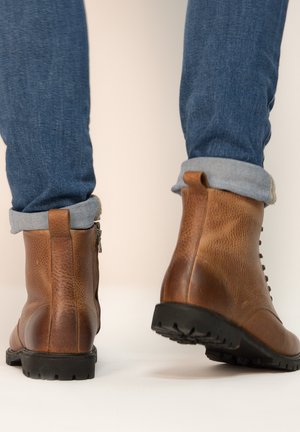 Botas de tobillo de cuero marrón con acabado texturizado, suelas de goma negra y diseño de cordones. Jeans con puños enrollados complementan las botas.
