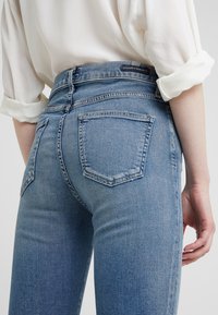 Jeans de mezclilla azul de tiro alto con acabado deslavado, dos bolsillos traseros y un ligero estiramiento. La cintura presenta una etiqueta de la marca en la parte trasera.