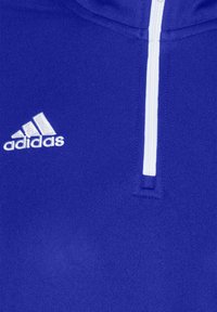 Gros plan d'un maillot de sport bleu avec une fermeture éclair blanche et un logo Adidas blanc brodé sur le côté gauche de la poitrine.