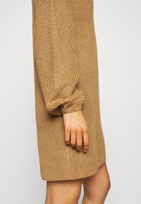 Stickad klänning i ljus tan, med ett strukturerat mönster, långa ärmar med ribbade manschetter och en avslappnad passform.