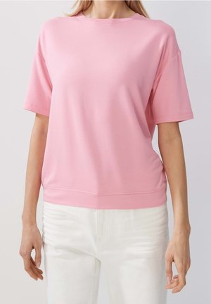 Kobieta ubrana w gładki różowy T-shirt z krótkim rękawem i białe spodnie stoi na tle jednolitego jasnego tła.