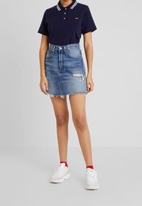 Personne portant un polo bleu marine, une jupe en jean taille haute usée, des chaussettes rouges aux chevilles et des baskets blanches, debout devant un fond uni.