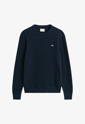 Granatowy sweter z dzianiny z teksturowanym wzorem, okrągły dekolt i małym logo na klatce piersiowej. Długie rękawy oraz ściągacze przy mankietach i u dołu.