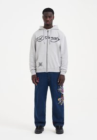 Gray zip-up hoodie with logo text. Blue denim pants featuring colorful dragon embroidery. Black sneakers complete the outfit.