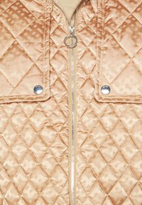 Beige gewatteerd jack met diamanten stiksel, voorrits met ronde trekker en twee klepzakken met glanzende zilveren knopen.