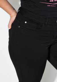 Musta farkut, joissa on istuva muotoilu ja jotka on valmistettu pehmeästä denim-kankaasta. Niissä on etutaskut ja metalliset yksityiskohdat.