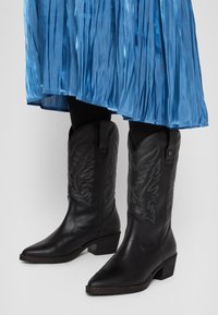 Botas vaqueras de cuero negro con punteras afiladas, diseños bordados en el caña y tacones de bloque. Combinadas con una falda azul con pliegues.