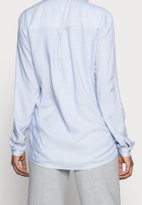 Lyseblå langærmet skjorte i blødt stof med rygplissé og knapmanchetter. Kombineret med lysegrå bukser.