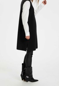 Schwarzes ärmelloses Kleid mit seitlichen Schlitzen, getragen über einem weißen Langarmhemd, kombiniert mit eng anliegenden schwarzen Hosen und spitzen schwarzen Ankle Boots.