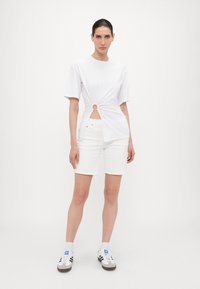 Hvit T-skjorte med knyttet vri og sirkulært utskjæring-detalj i midjen, kombinert med hvite shorts og uformelt fottøy. Jevn tekstur.