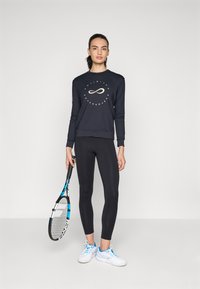 Marineblå hettegenser med grafikk som sier "uendelig ytelse", kombinert med svarte leggings. Holder en blå og svart tenniskølle, iført hvite sko.