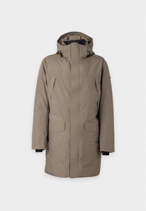 Homme portant un manteau d'hiver à capuche taupe, pantalon gris clair et baskets blanches, debout devant un fond clair uni.