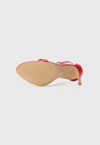 Sandale à talon haut rouge avec un design élégant, un dessus en synthétique lisse, un bout pointu et une semelle beige texturée avec un petit accent doré sur le talon.