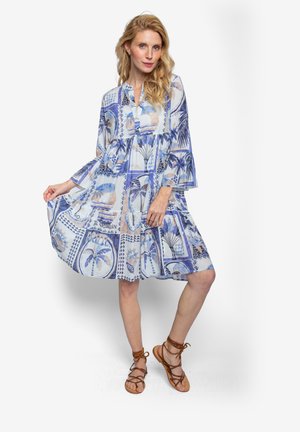 Leichtes, transparentes Kleid in Blau mit Palmen- und geometrischem Muster. Mit Glockenärmeln und gestuftem Saum. Kombiniert mit braunen Sandalen.