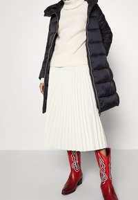 Sort puffershirt med hætte, hvid ribbet sweater, plisseret hvid nederdel og røde cowboystøvler med sort detaljer. Glatte teksturer, varierede former.