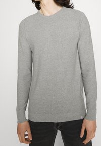 Un pull gris clair à manches longues en tissu doux, avec un col rond et des poignets côtelés, porté avec un jean foncé.