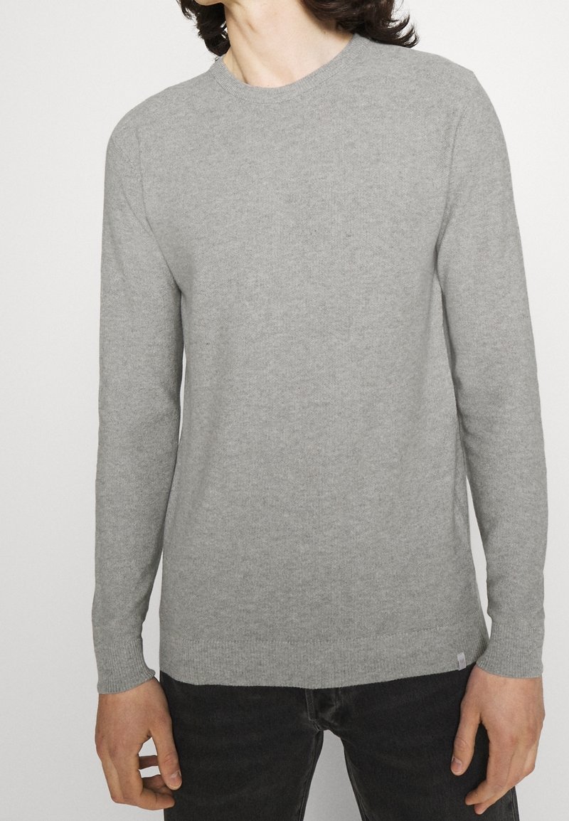 Un pull gris clair à manches longues en tissu doux, avec un col rond et des poignets côtelés, porté avec un jean foncé.