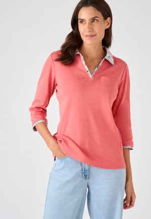 Vrouw draagt een koraalroze shirt met lange mouwen met bloemenkraag en manchetten, lichtblauwe spijkerbroek, hand in de zak, kijkt opzij.