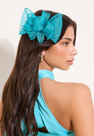 Femme aux longs cheveux bruns portant un haut turquoise sans manches et un grand nœud en résille turquoise dans les cheveux, regardant vers la droite.