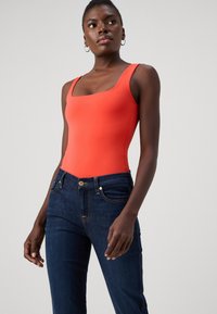 Κόκκινο κοντομάνικο κορμάκι με τετράγωνη λαιμόκοψη, συνδυασμένο με σκούρες μπλε τζιν παντελόνες που έχουν χρυσές λεπτομέρειες και κίτρινη ραφή στις τσέπες.