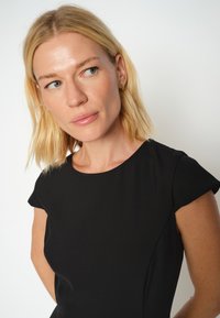 Rochie neagră cu decolteu rotund și mâneci scurte tip capă. Material neted, mat, cu o siluetă ajustată. Detalii minore de cusătură pe laterale.