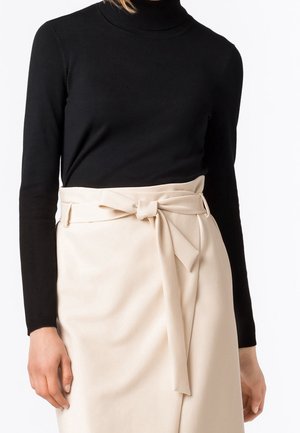 Personne portant un col roulé noir à manches longues et une jupe crème taille haute attachée avec une ceinture en tissu nouée en nœud.