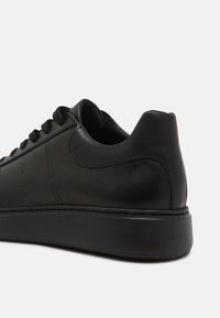 Svart lädersneaker med slät yta, rund tå och flat sula. Har texturerade detaljer och svarta skosnören.