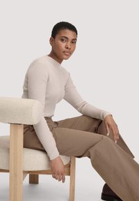 Strakke ribbelige lichtbeige trui met hoge hals, gedragen met ontspannen tankleurige broeken, zittend op een licht houten stoel met een gewatteerde zitting.