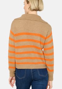 Khaki pleteno pulover s oranžnimi vodoravnimi črtami, ki ima zloženi ovratnik in žepaste manšete, nosen z modrimi džinsom.