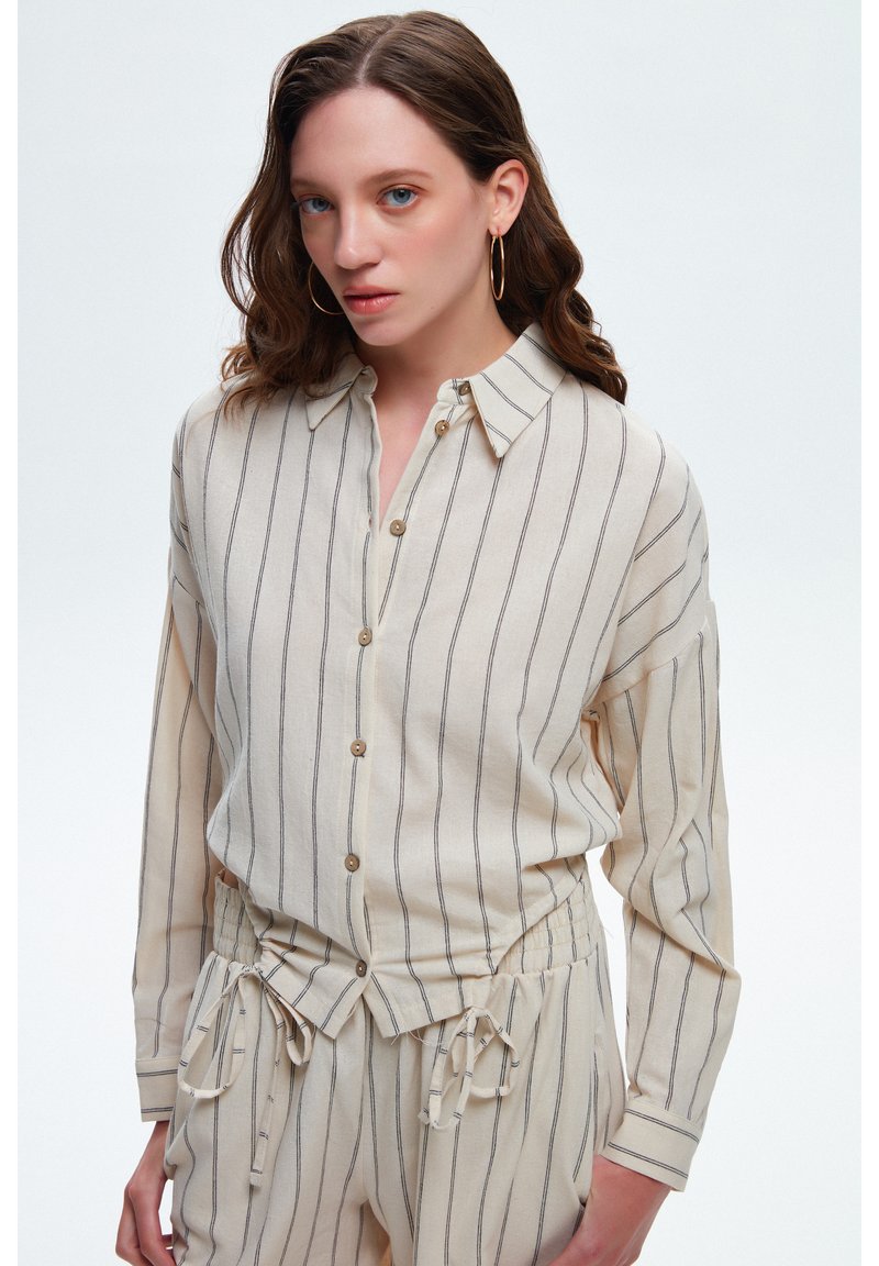 Camicia button-up beige chiaro con righe verticali blu navy, dotata di colletto e vestibilità comoda. Indossata con pantaloni abbinati con coulisse.
