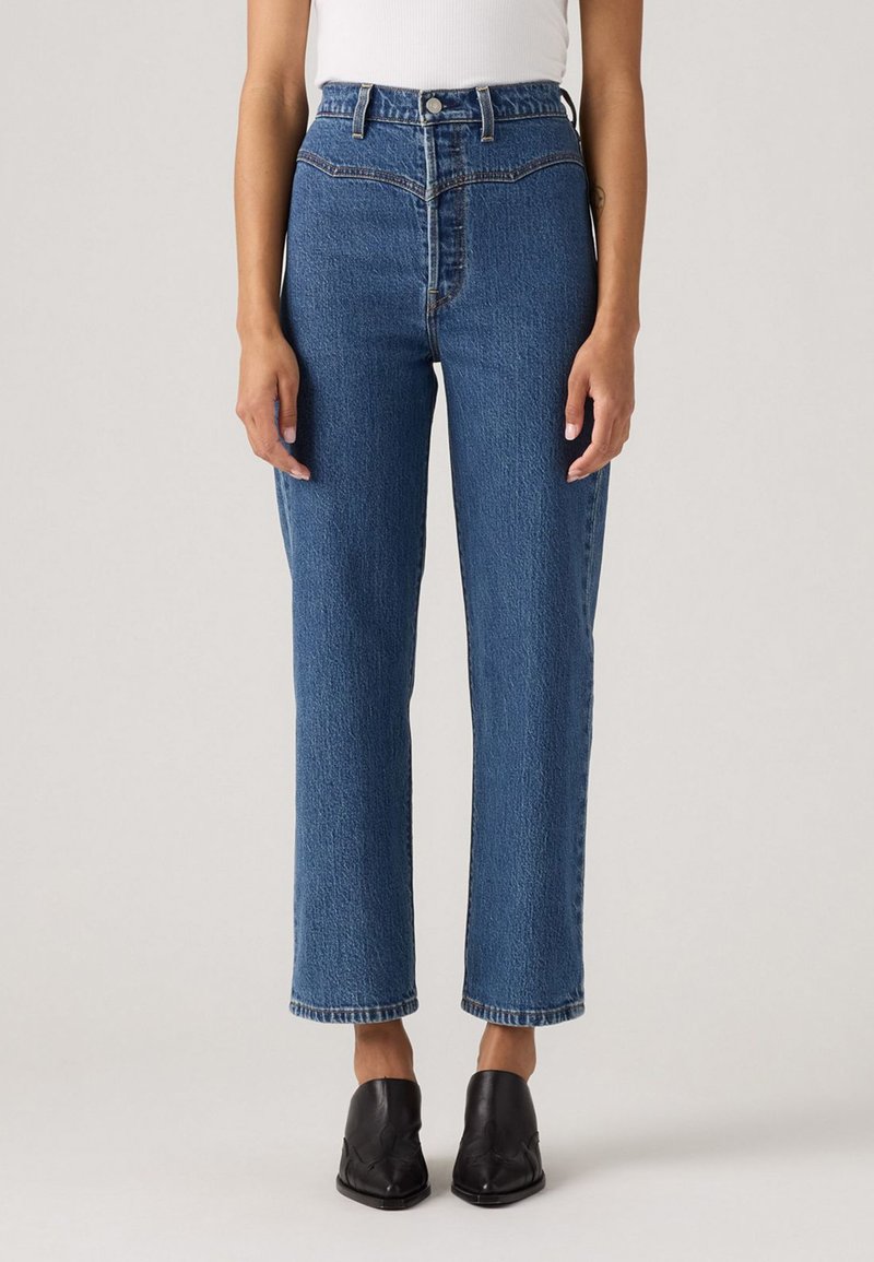 Donna che indossa jeans blu a vita alta e dritti, con scarpe nere a punta, in piedi davanti a uno sfondo neutro.