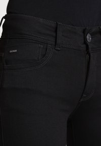 Jeans de mezclilla negros con un diseño ajustado, que presentan un cierre de botón de metal, un pequeño logo "RAW" y detalles de costura en contraste.